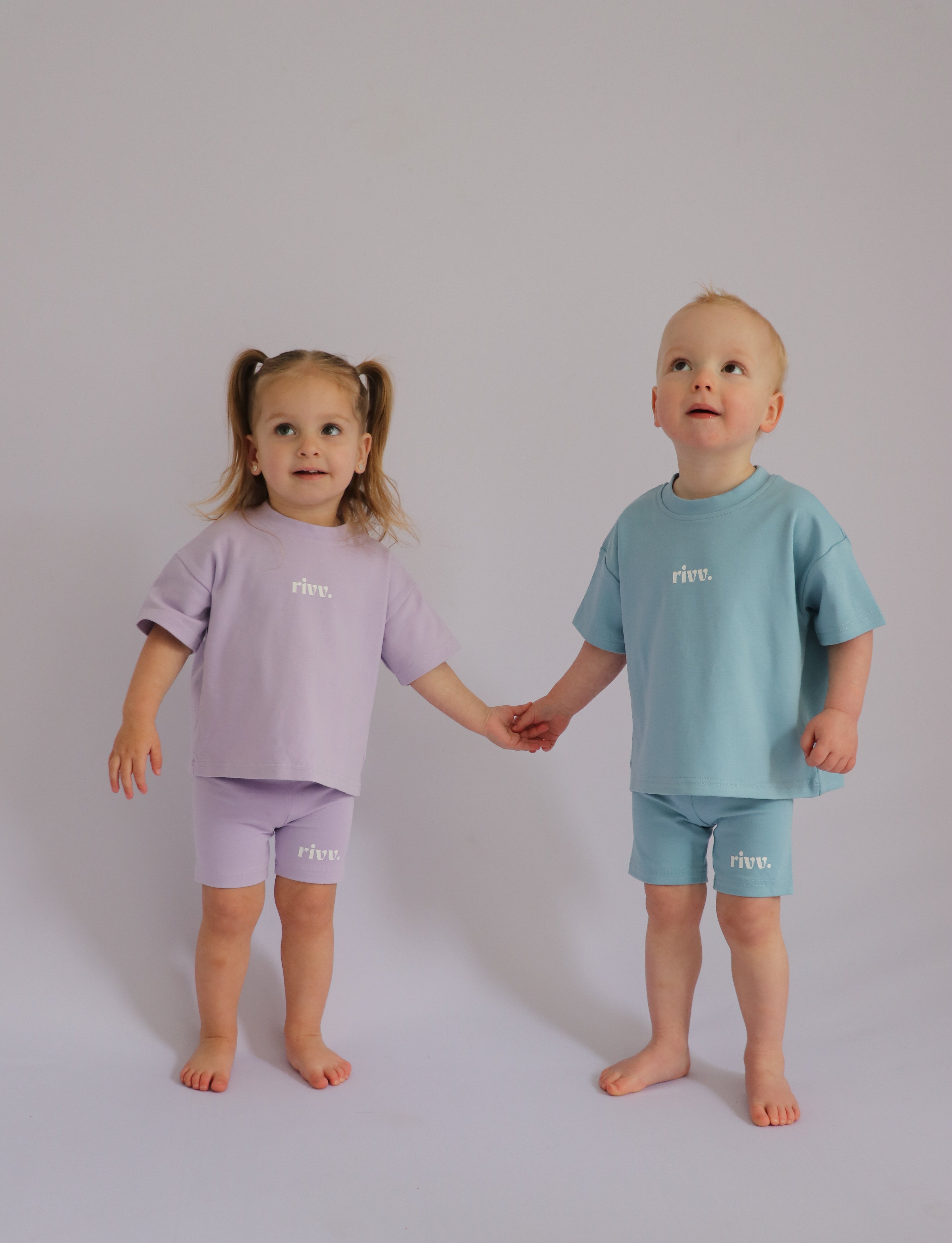 Two children holding hands, wearing matching tee sets with a brand logo on a plain background.