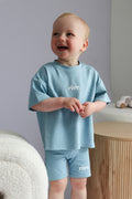 Child wearing a light blue 'rivv ' t-shirt and shorts standing in a room with a neutral background.
