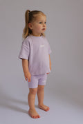 Young child wearing a light purple outfit with visible branding on a plain background