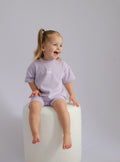 Child wearing a light purple outfit sitting on a white stool against a plain background