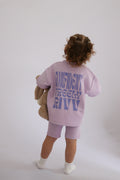 Child wearing a lavender outfit with text on the back, holding a teddy bear against a white background