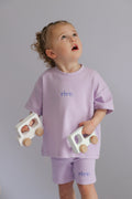 Child wearing a lavender outfit with 'Rivv ' branding, holding wooden toys against a gray background