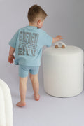 Child wearing a light blue outfit with text, standing next to a white ottoman.