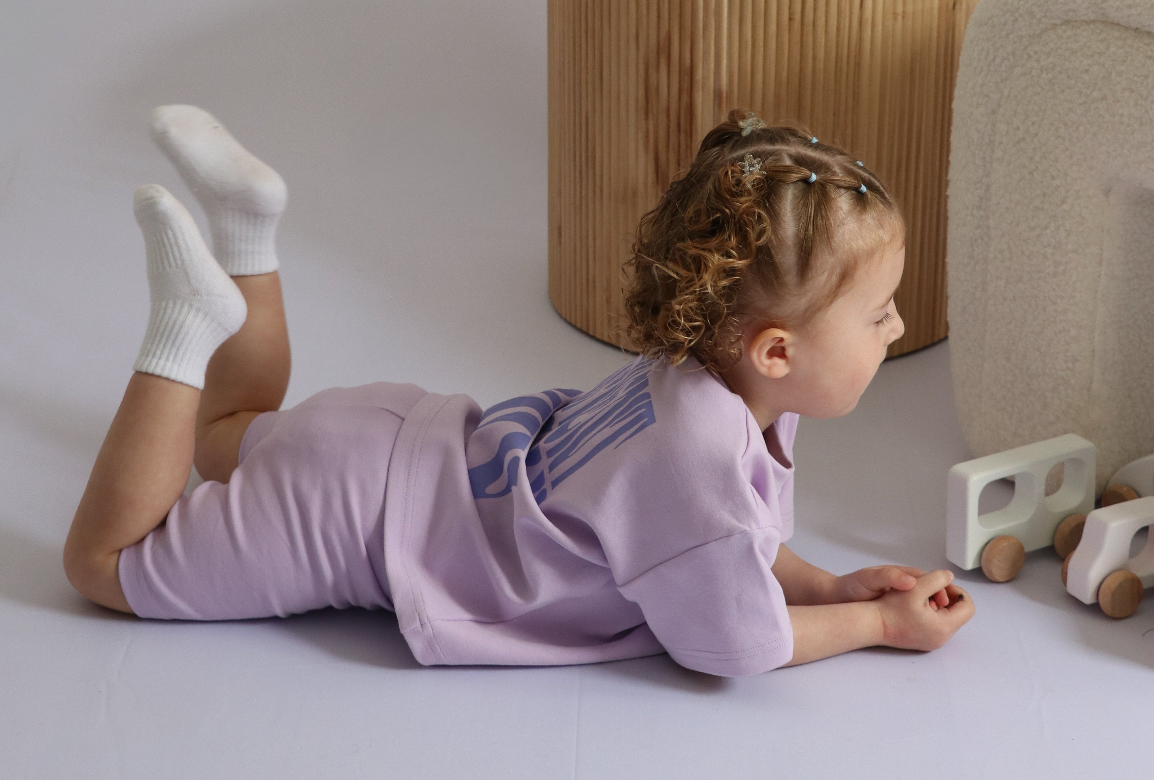 Child in a purple outfit lying on a white floor with a wooden stool and toy in the background