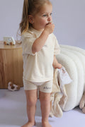 Child wearing a Beige t-shirt and shorts with 'Rivv' branding, standing indoors.