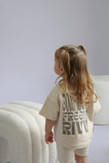 Child wearing a beige outfit with text, standing in a minimalistic room.
